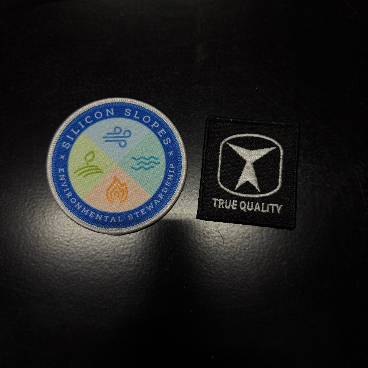 Two patches on a dark surface, one labeled 'Silicon Slopes Environmental Stewardship' and the other 'True Quality'.