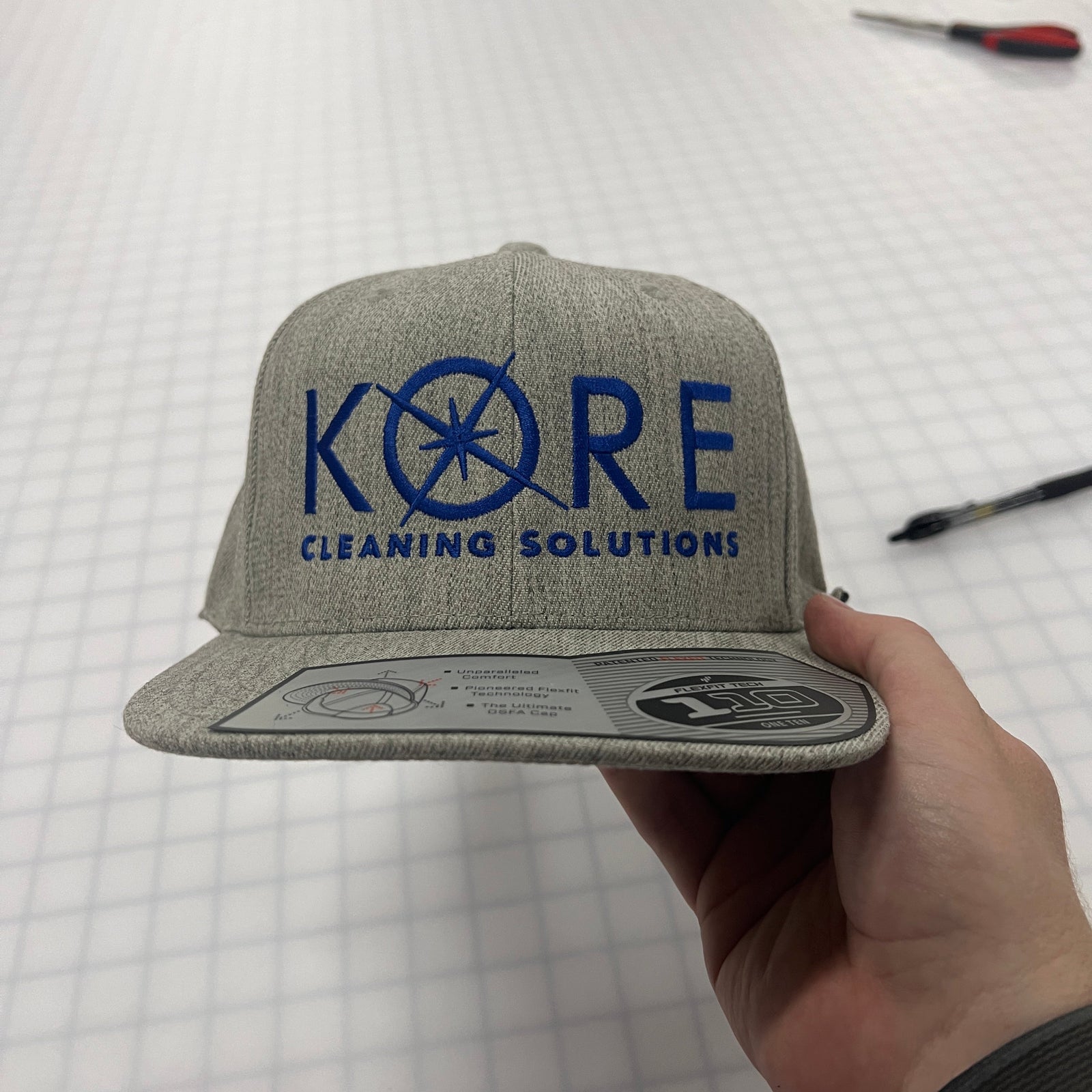 Gray cap with blue 'Kore Cleaning Solutions' logo held by a hand on a white background.