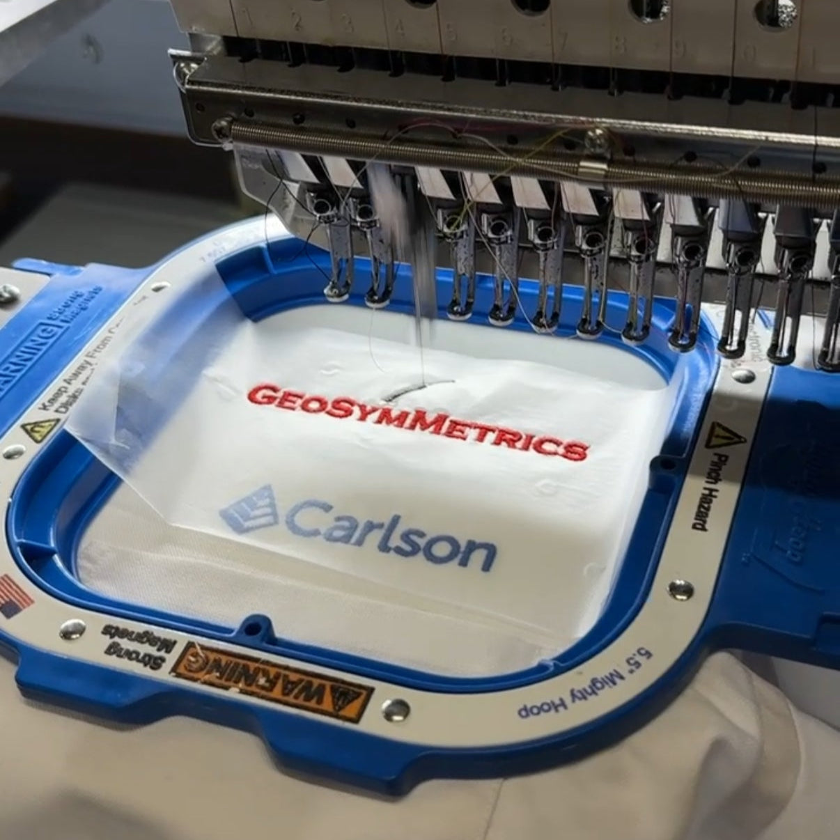 Embroidery machine with a white fabric sample featuring 'Geosymmetrics' and 'Carlson' logos.